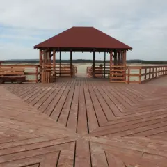 Drewniana promenada prowadzi do altany na pustyni błędowskiej. W tle widać piaszczysty teren i drzewa.