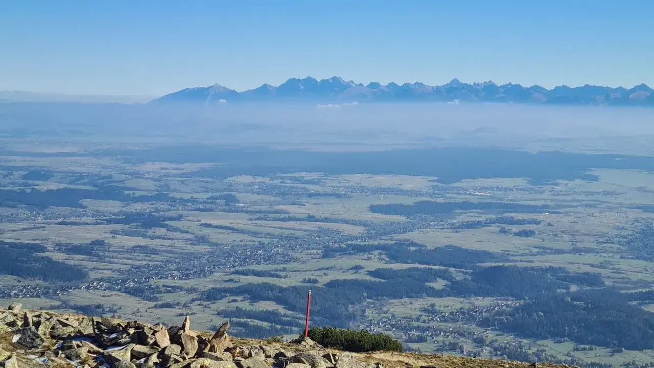 Widok z Babiej Góry na Tatry. Mgła spowija doliny, a szczyty górskie rysują się na tle błękitnego nieba.